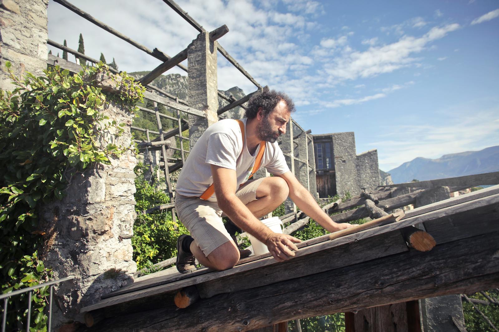 A man kneeling on a rooftop under construction in a picturesque mountainous landscape on a sunny day.